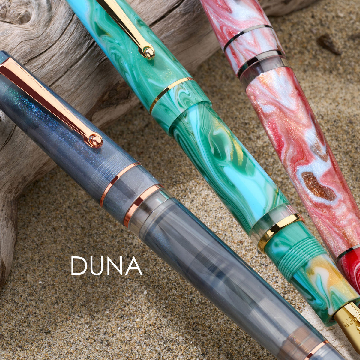 Three artistic pens with marbled designs on a textured surface, featuring the brand Delta 'DUNA'.